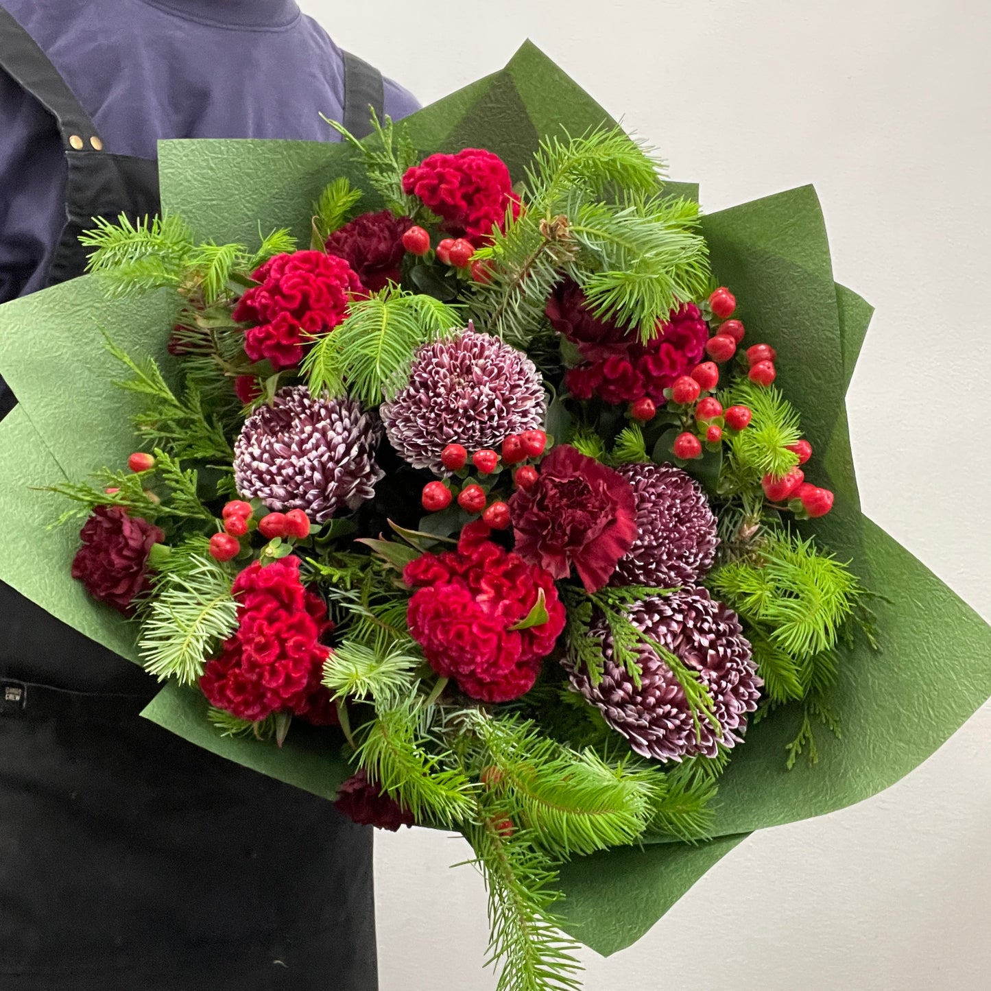 Christmas bouquet featuring red and burgundy celosia, hypericum berries, disbuds, and seasonal Christmas foliage, wrapped for the holiday season.