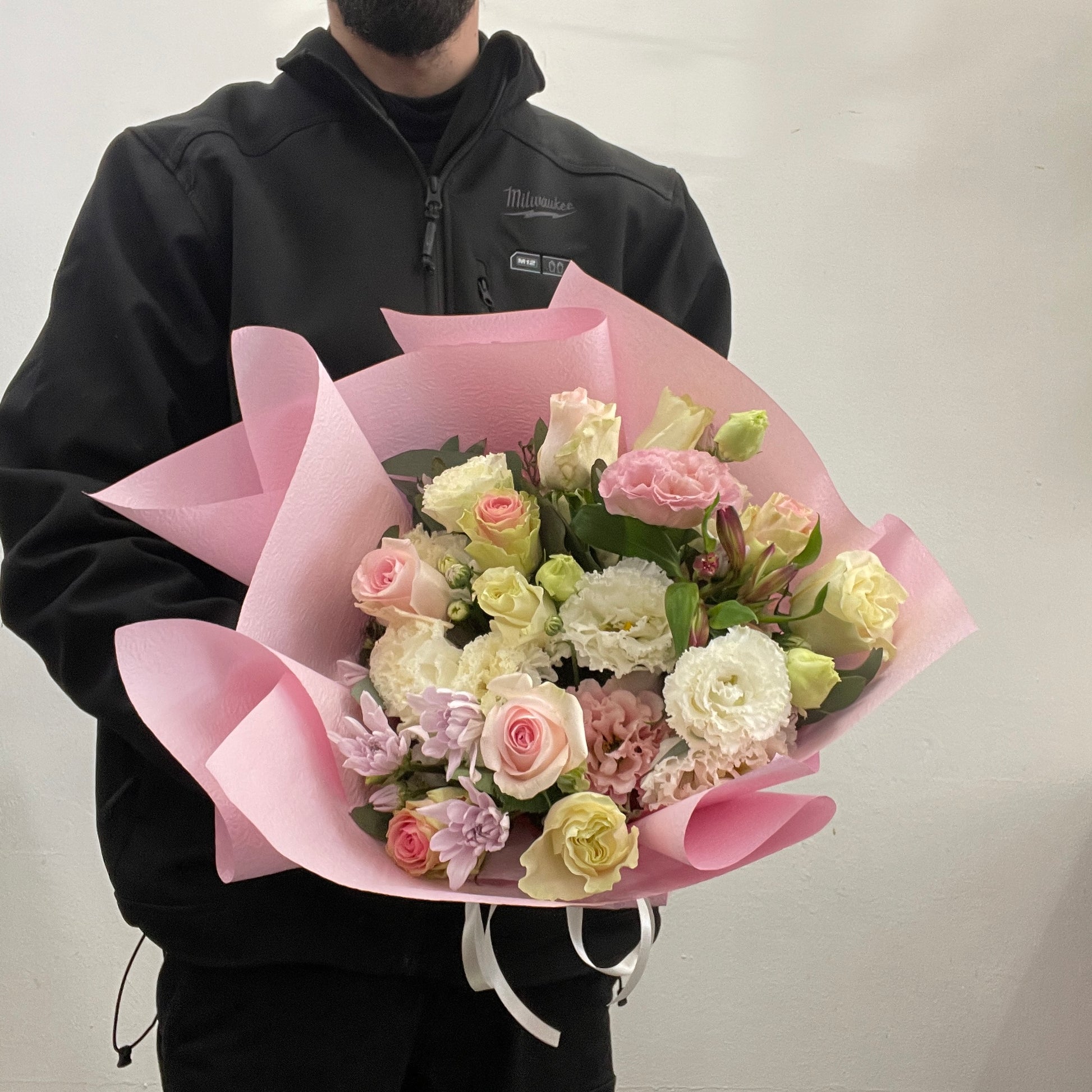 Person holding a arrangement  of flowers wrapped in pink paper against a plain background