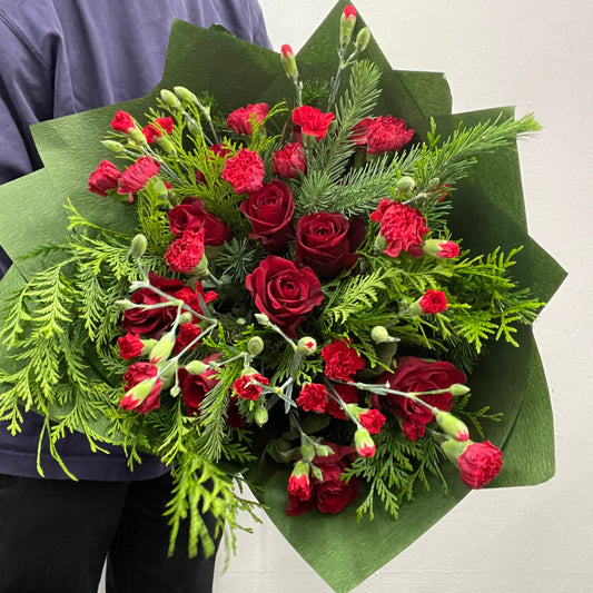 Red and green Christmas bouquet featuring red roses and festive foliage, photographed as a hand-tied arrangement.