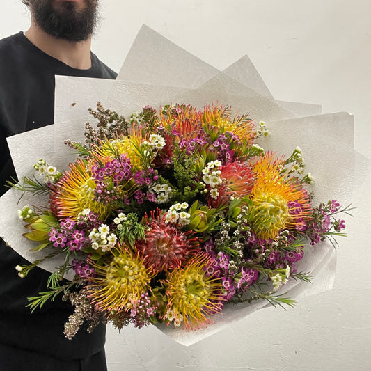 Rounded bouquet featuring native pincushions in orange and yellow tones with colourful foliage, part of the Spring Limited Collection 2025 by Adam The Flower Man.
